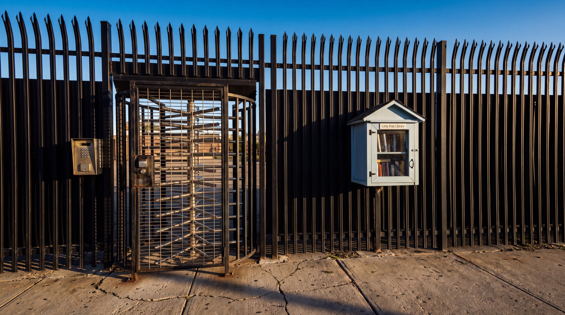 A tall black steel anti-climb security fence with curved spear tops, rendered in low golden light. A full-height interleaved steel exit turnstile sits caged in the fence on the left, with an intercom keypad beside it. Mounted further along the fence, small against it, is a white Little Free Library box with a glass door and two shelves of books. Cracked concrete underfoot, weeds in the seams, deep blue sky above.
